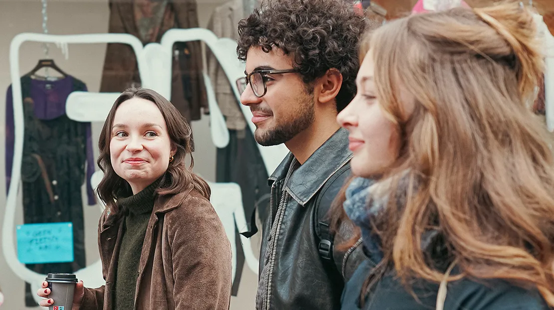 Diverse group of three Webster Leiden students walking outdoors past local shopfronts, one holding a takeaway coffee.