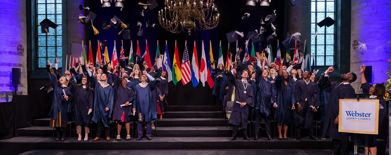 Group of Leiden grads throwing hats in the air with international flags in the background. 